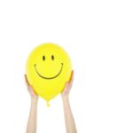 Cheerful yellow balloon with a smiley face held up by hands against a white background.